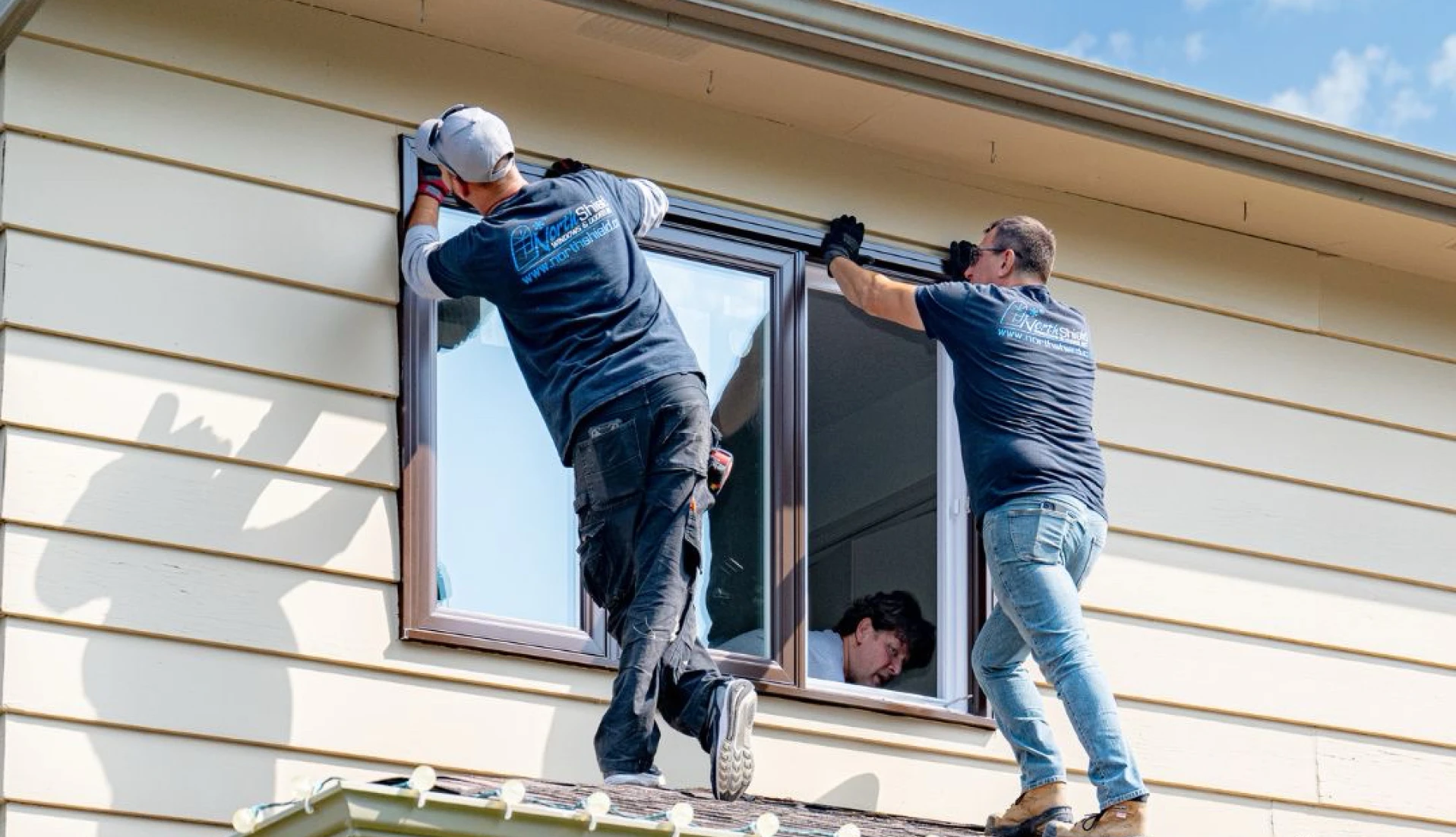 Three workers installing a new window on the second story of a beige house with a brown trim and gutter